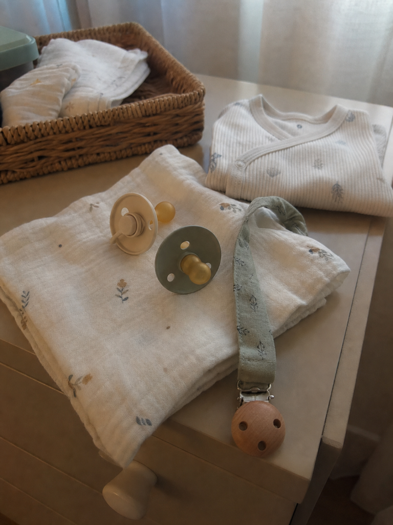Chupetas e porta-chupeta sobre musselina numa cómoda de bebé com luz suave